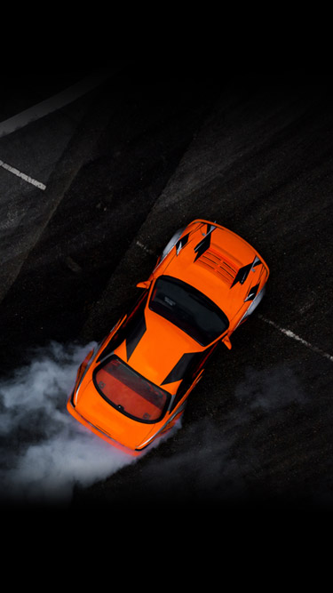 An aerial shot of an orange race car making a sharp turn on a black road with smoke coming out the back